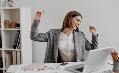 Positive office worker in white shirt dancing while sitting at table on of shelves with documents.