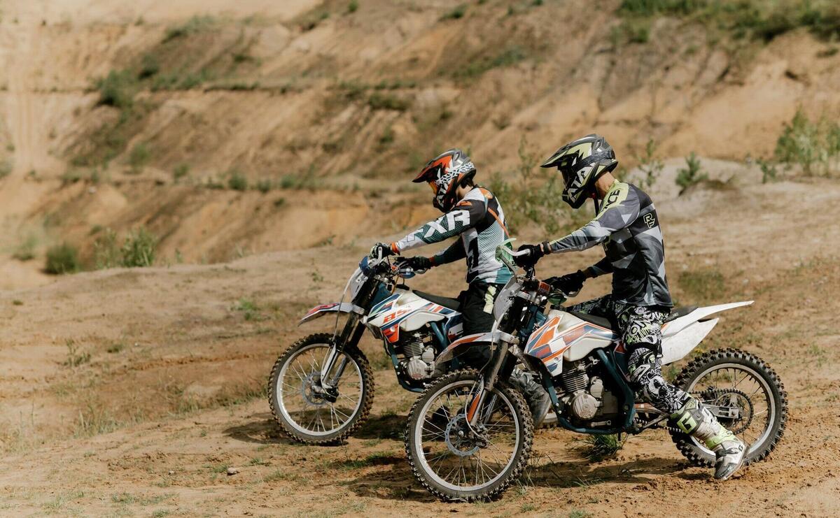 Two dirt bikers equipped with helmets prepare for a ride on a sandy outdoor trail. Enthusiastic and energetic.