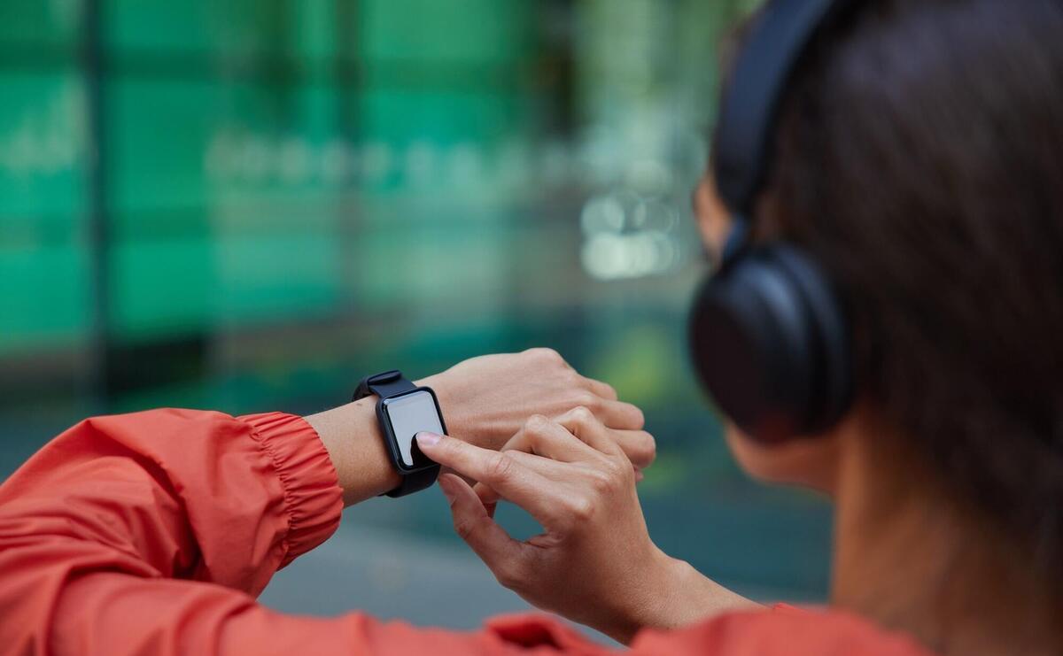  woman checks results of fitness training on smartwatch listens music via headphones dressed in anorak poses on blurred 