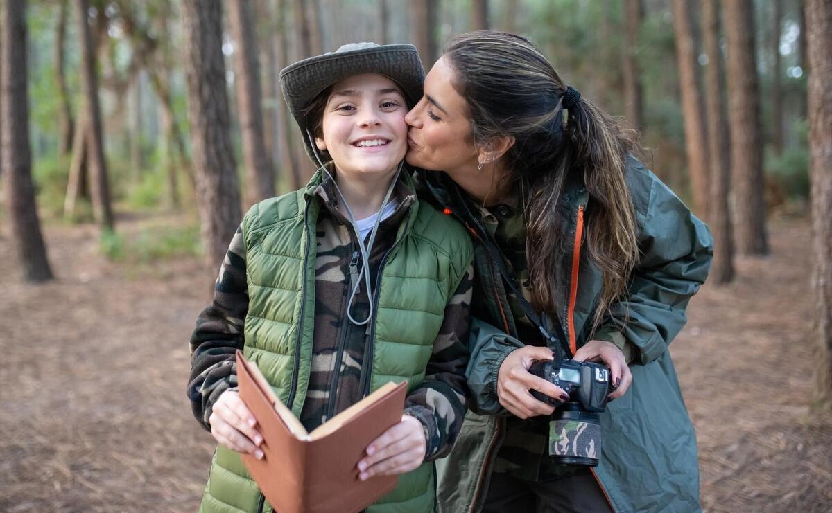 Tender mother with son with camera in forest. Female model in sportive clothes kissing smiling boy on cheek. Hobby, photography concept