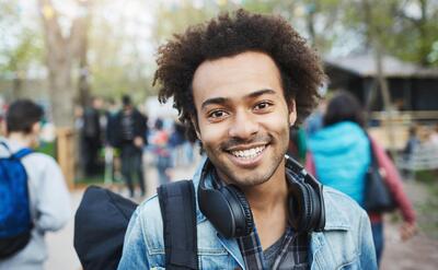 Close-up shot of happy emotive young african-american guy with afro hairstyle and bristle, smiling broadly while wearing denim coat and backpack, walking across park during festival