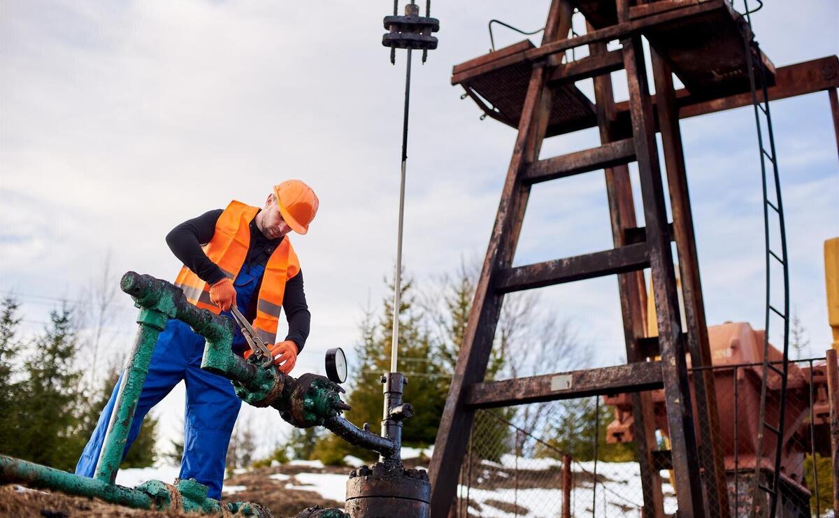 Oil worker in orange uniform and helmet working with a pipe wrench near an oil pump jack