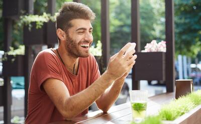 Handsome happy guy sitting in cafe, drinking lemonade and using mobile phone, laughing over text message