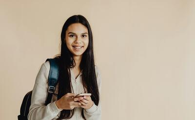 Teen girl with backpack using a smartphone against a beige background, smiling.