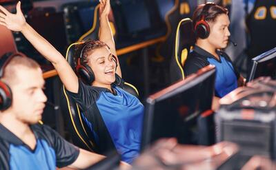 Young excited mixed race girl, female cybersport gamer wearing headphones raising hands up, celebrating success while participating in eSport tournament, playing online video games professionally