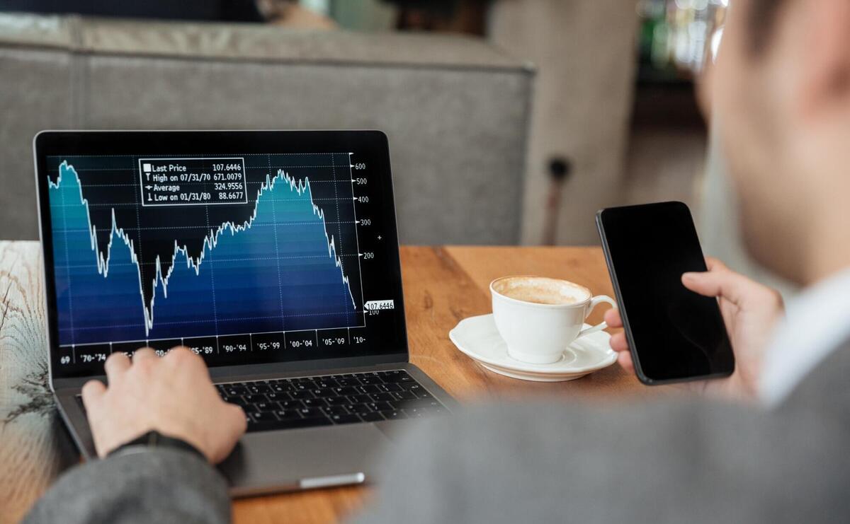 Cropped image of businessman sitting by the table in cafe and analyzing indicators on laptop computer while using smartphone