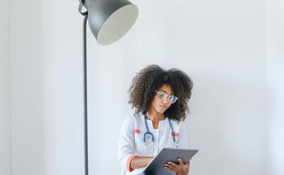 A healthcare worker with curly hair and eyeglasses using a tablet next to a floor lamp.