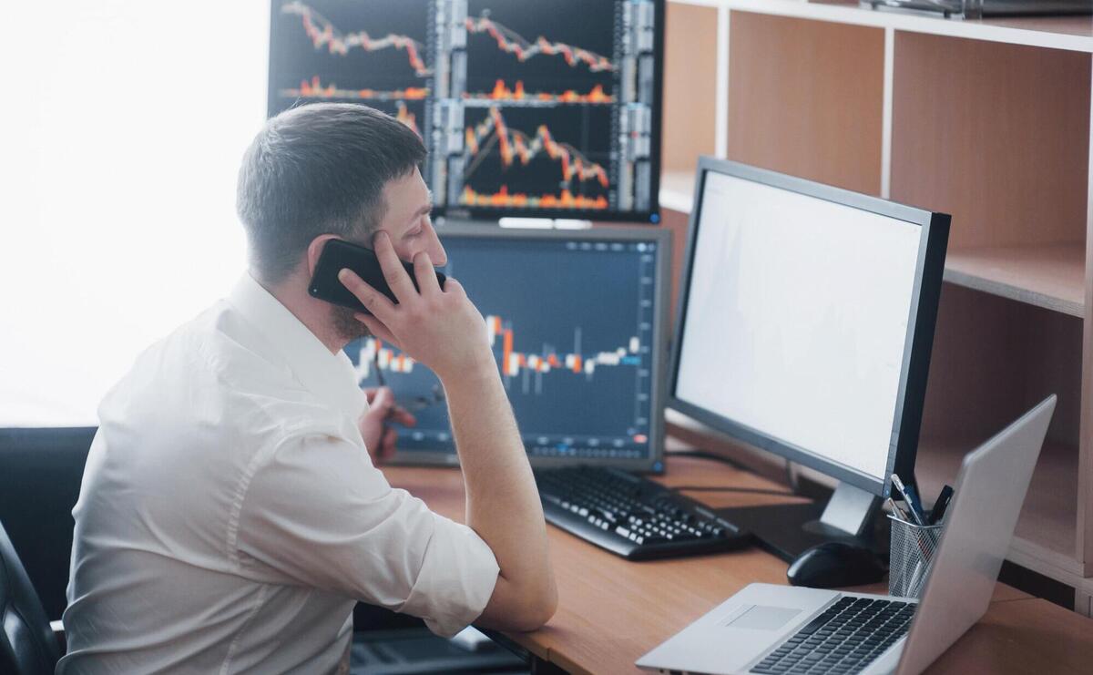 Over the shoulder view of and stock broker trading online while accepting orders by phone. Multiple computer screens ful of charts and data analyses in background