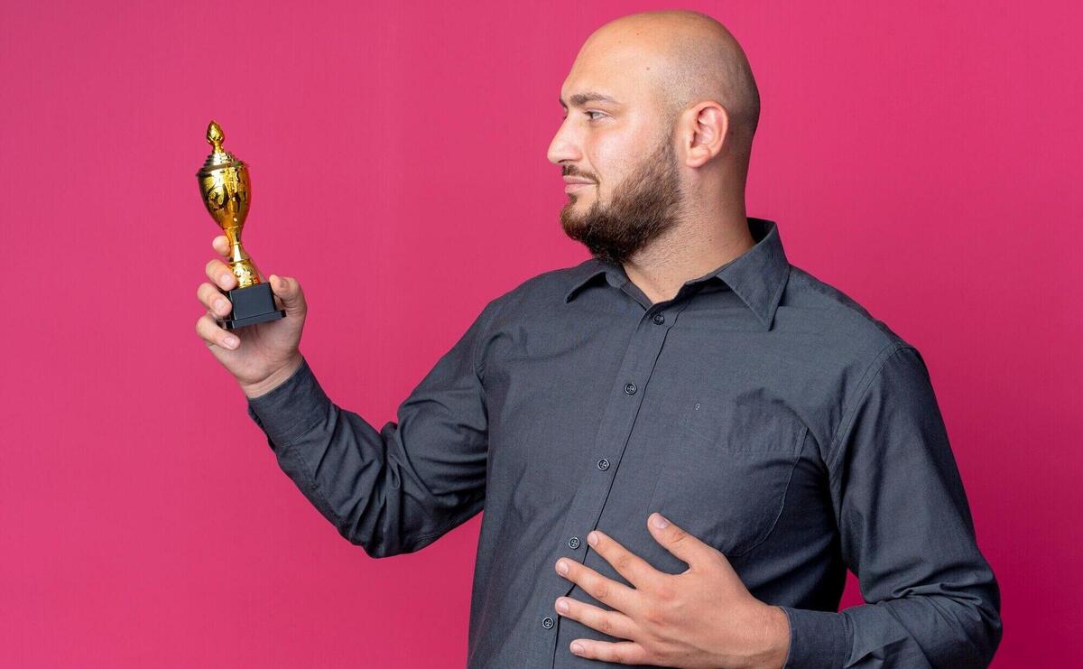 Pleased young bald call center man holding and looking at winner cup and putting hand on belly isolated on crimson background