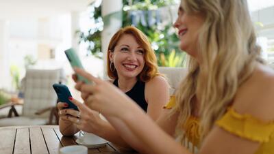 Two women using smartphones and drinking coffee sitting on table at home terrace