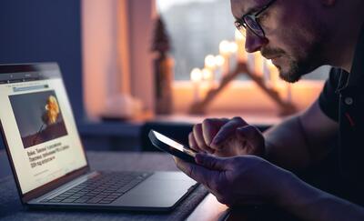 A man with a smartphone sits in front of a laptop late at night