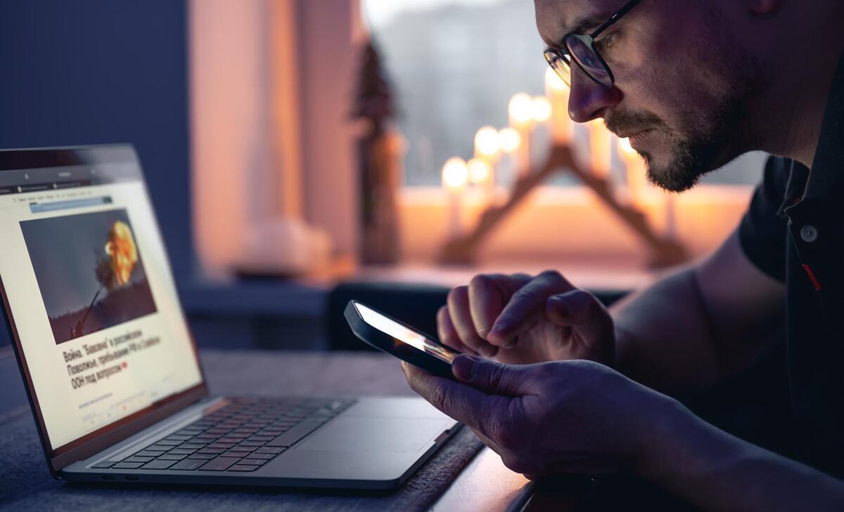 A man with a smartphone sits in front of a laptop late at night