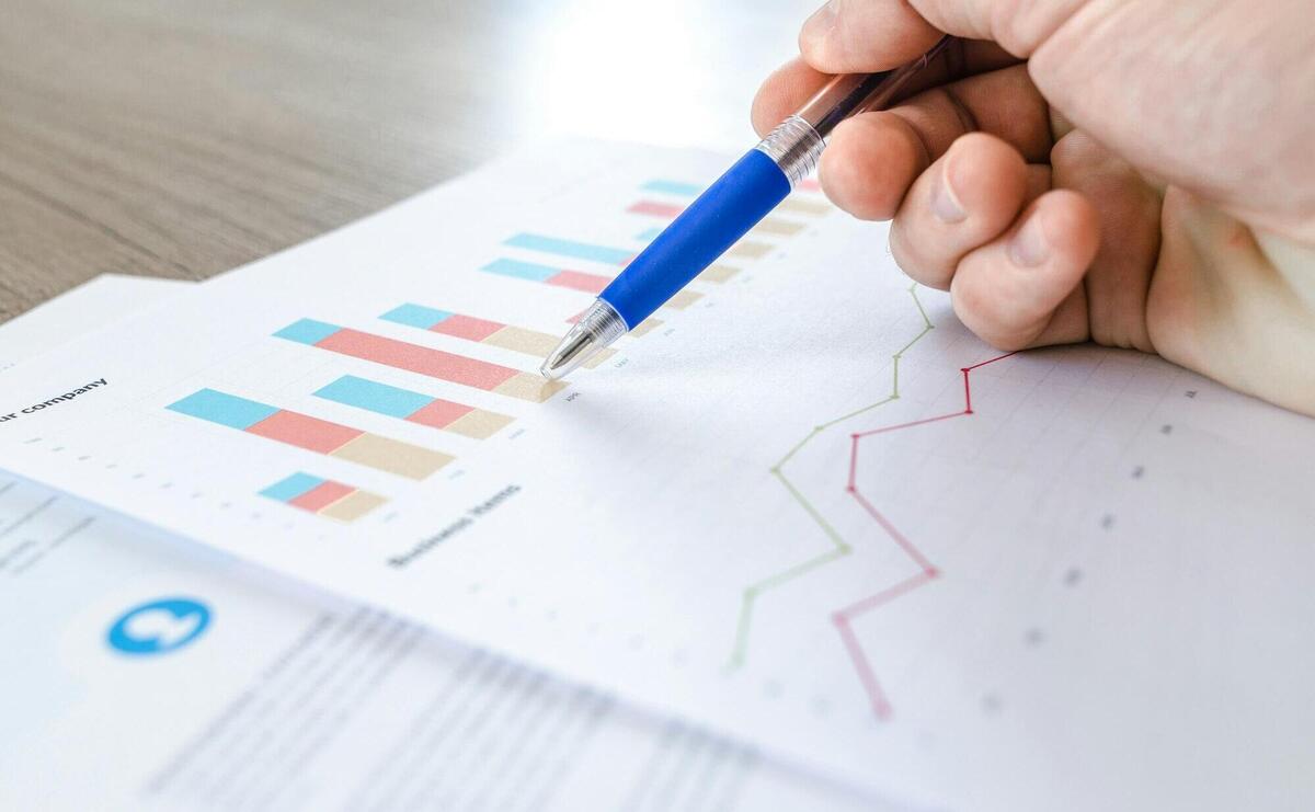 A close-up of a hand with a pen analyzing data on colorful bar and line charts on paper.