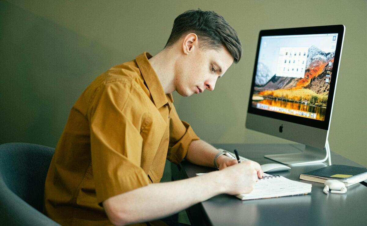 Adult male concentrating on notes with a desktop computer in a home office setting.