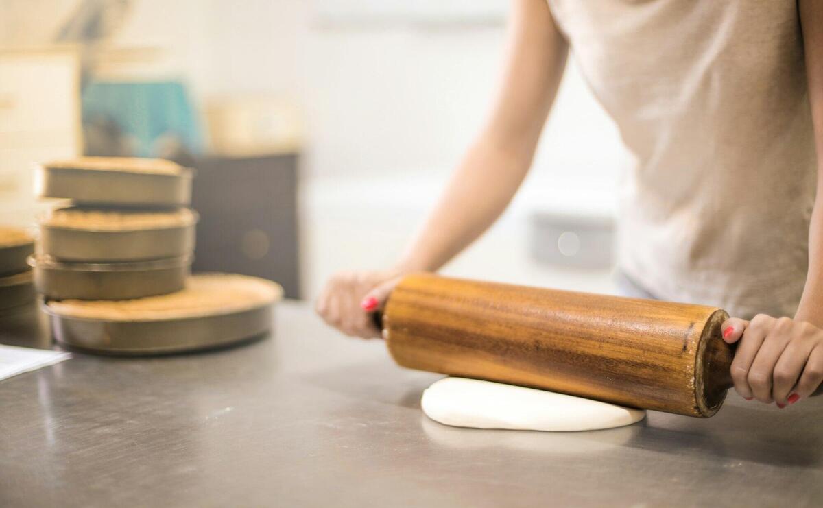 A woman using a wooden rolling pin to flatten dough in a bakery kitchen setting.