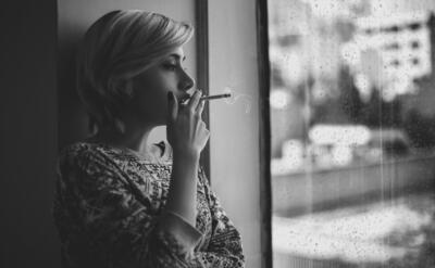 Black and white depressed female with short haircut smoking cigarette and looking out window at rainy city