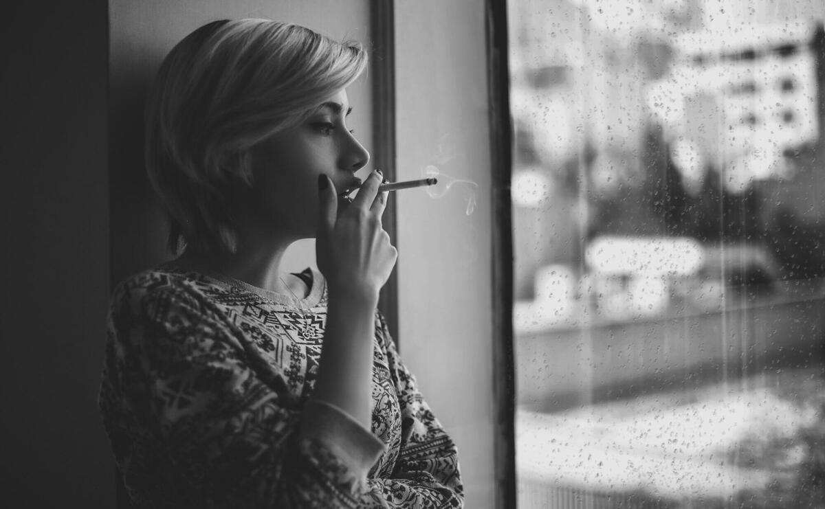 Black and white depressed female with short haircut smoking cigarette and looking out window at rainy city