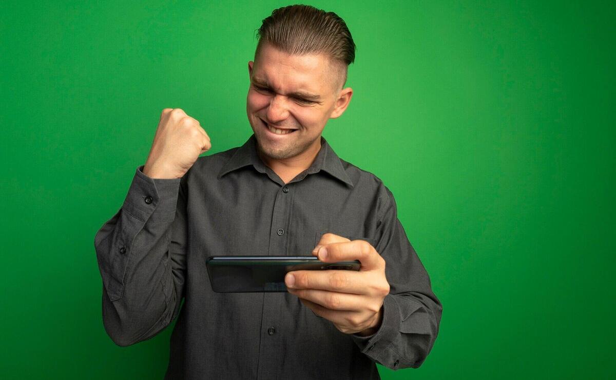 Young handsome man in grey shirt using gadget clenching fist happy and excited rejoicing his success standing over green wall