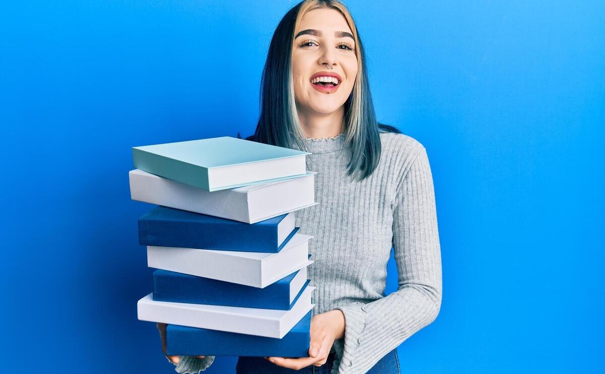 Young modern girl holding a pile of books smiling and laughing hard out loud because funny crazy joke