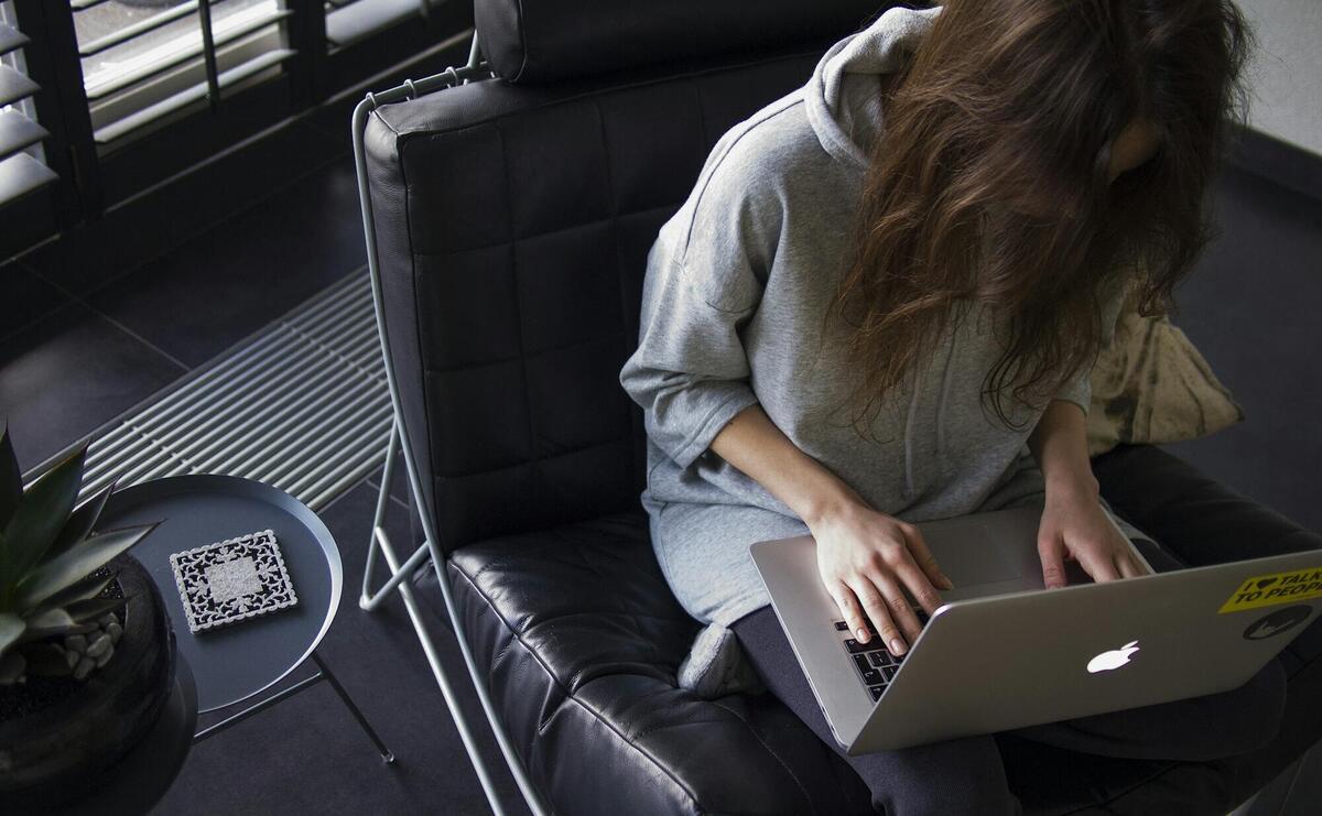 woman in blue chambray long-sleeved top sitting on black leather chair with silver MacBook on lap