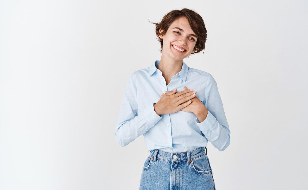 Portrait of smiling caucasian woman holding hands on heart and thanking you, feeling grateful, standing on white wall