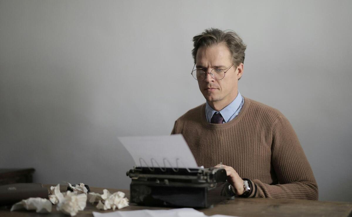 Man concentrating on writing at a desk with a vintage typewriter surrounded by crumpled paper.