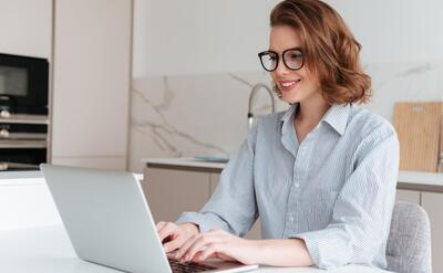 Elegant smiling woman in glasses and striped shirt using laptop computer while siting at table in kitchen