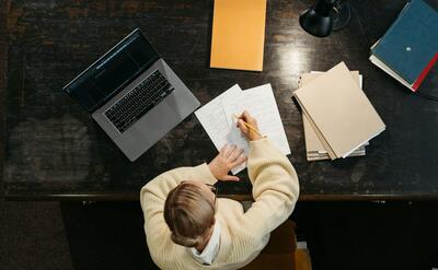 Overhead view of woman writing at desk with laptop, papers, and books.