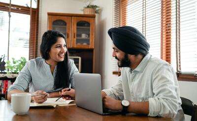 Laughing Indian woman with husband sitting at table and discussing project while working together with laptop and notebook at home