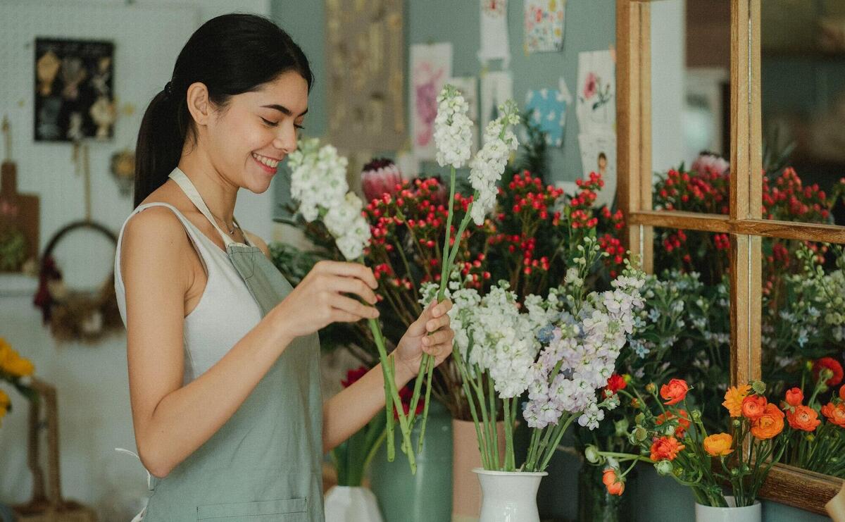 Side view of smiling florist with long dark hair in apron standing near counter and smelling flowers in vases in cozy floristry studio