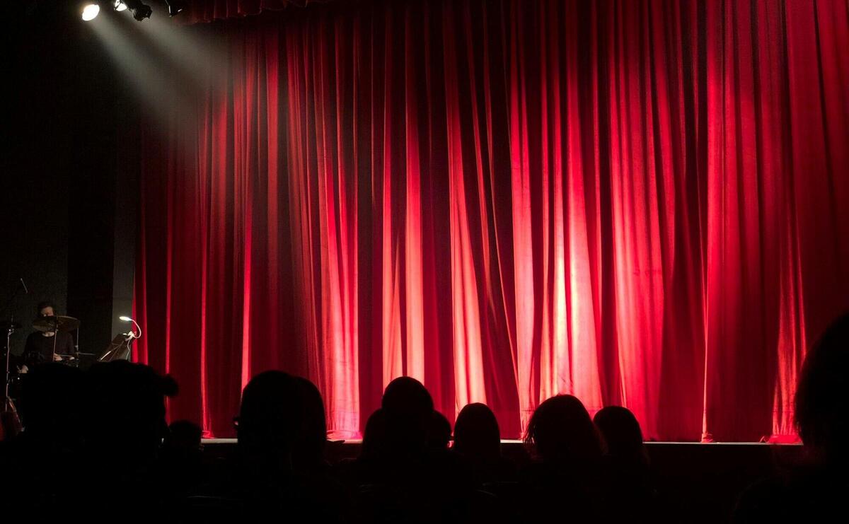 Dimly lit theater stage with red curtains and audience silhouettes under spotlights.