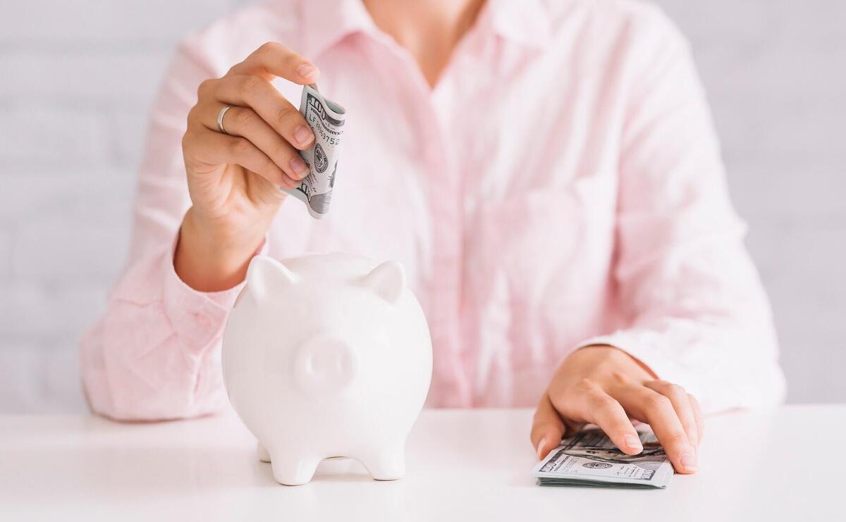 Close-up of woman inserting hundred dollar currency in white piggybank