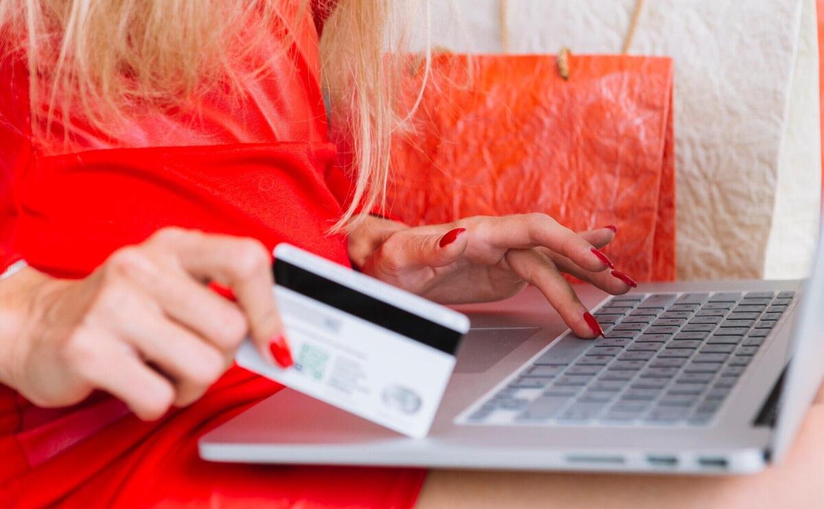 Woman in red sitting with laptop and card near shopping bags 