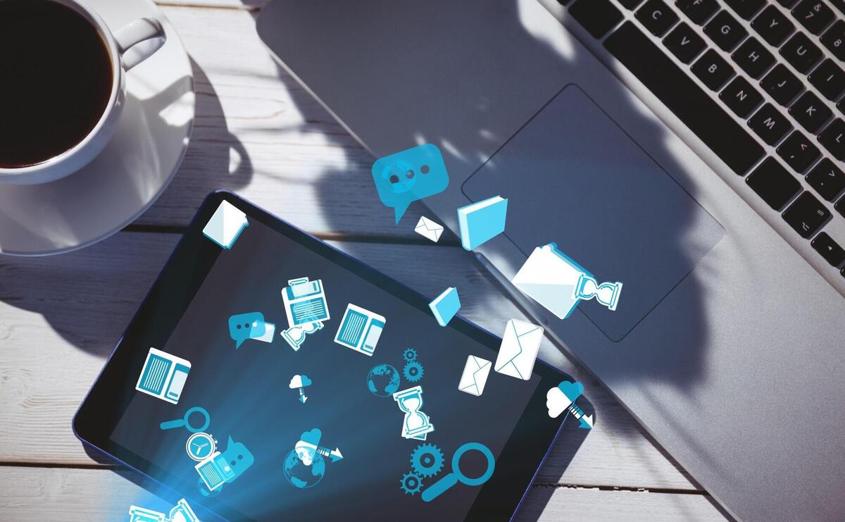 Tablet displaying digital icons beside a laptop and coffee on a wooden table.